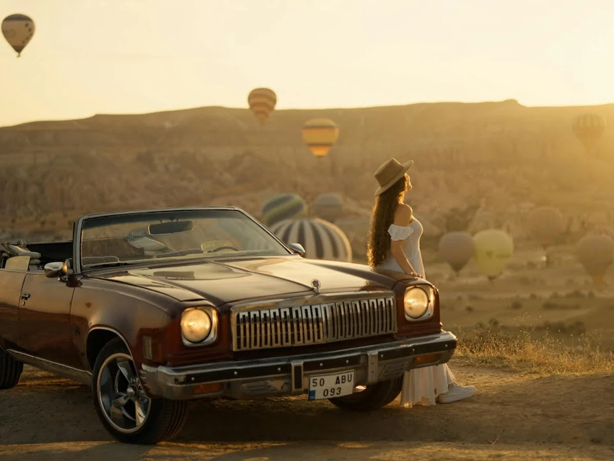 Black classic convertible with a woman posing at sunrise among Cappadocia balloons