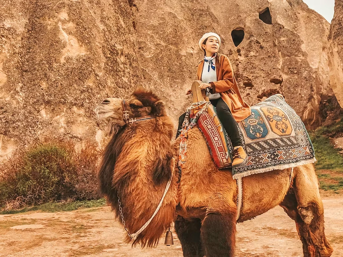 Woman posing on a camel beside Cappadocia cave formations during a daytime ride