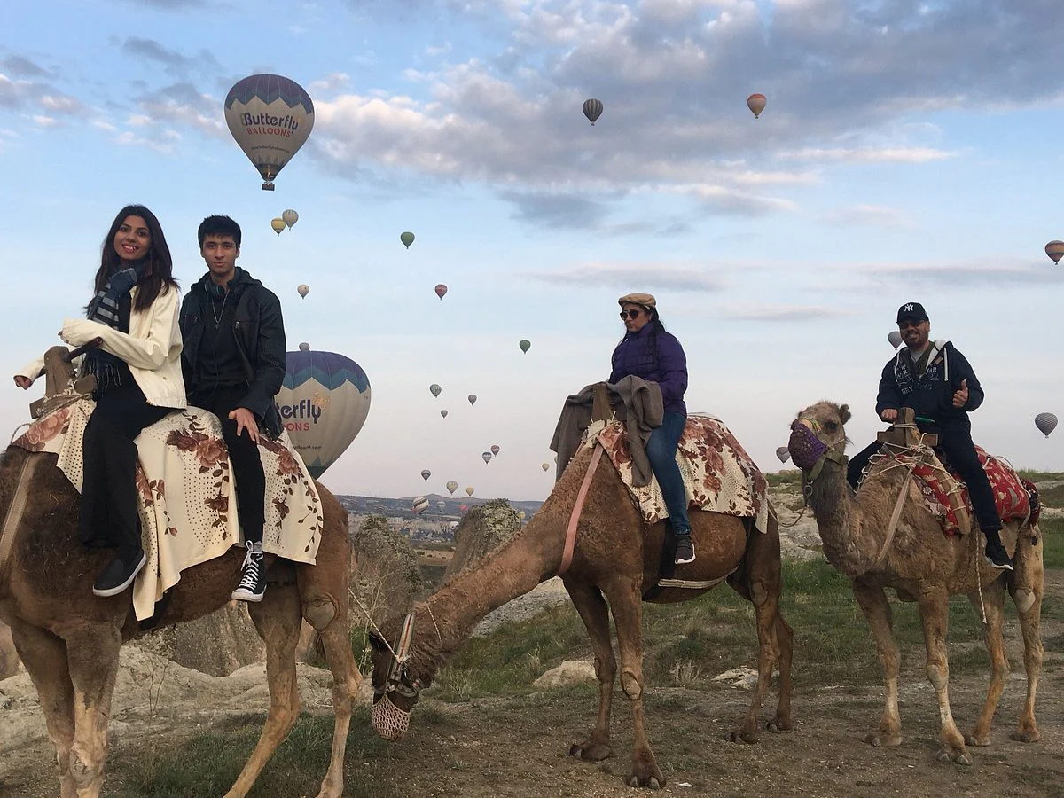Guests riding decorated camels through a Cappadocia valley with balloons overhead