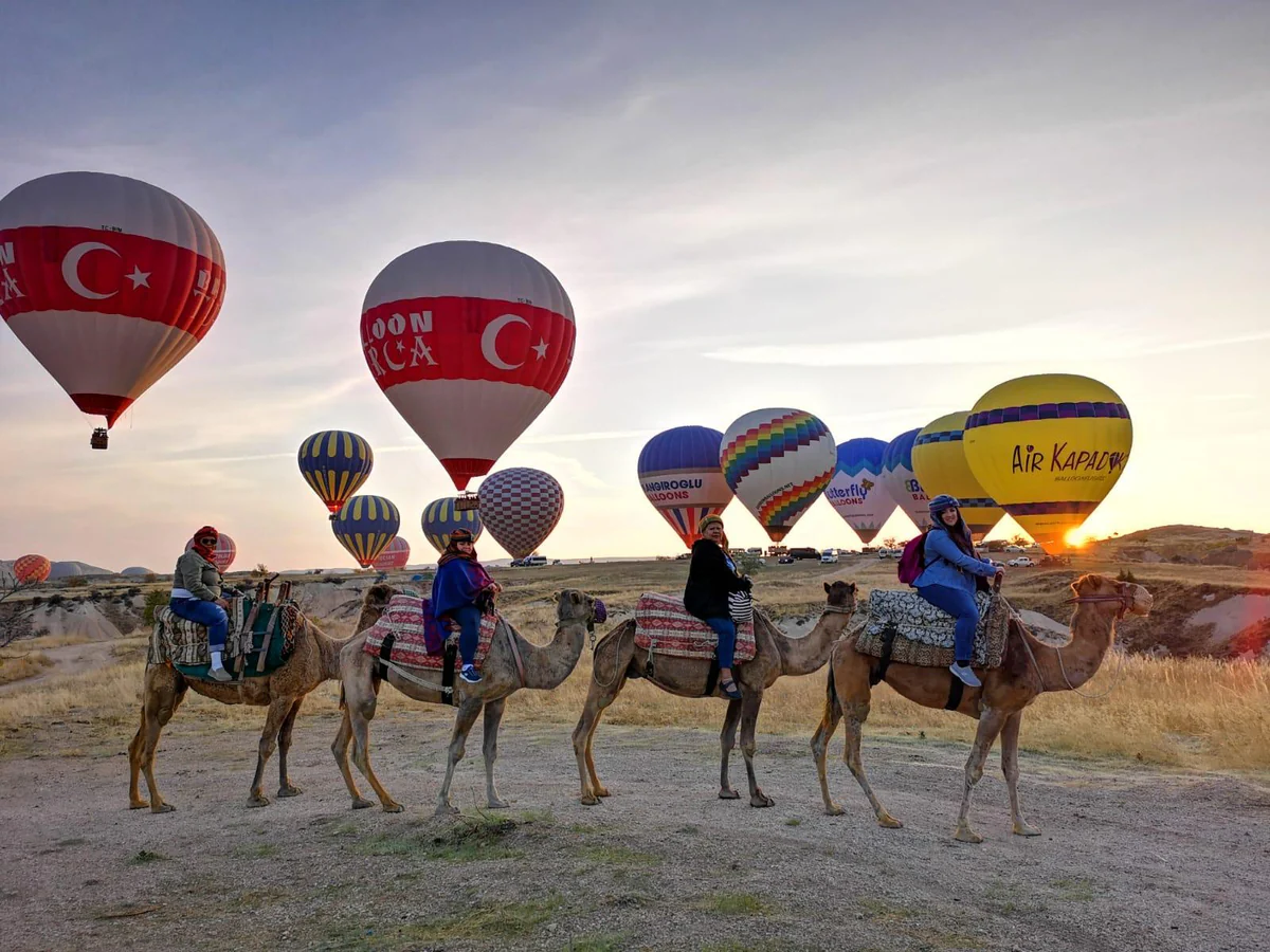 Camel safari group posing beneath hot air balloons in Cappadocia at sunrise