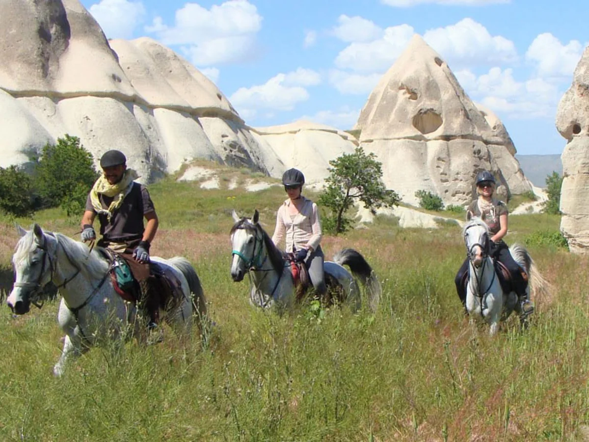 Travelers riding horses through green fields beside Cappadocia fairy chimneys
