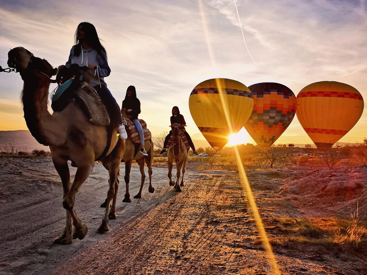 Camel riders passing hot air balloons during sunrise in Cappadocia