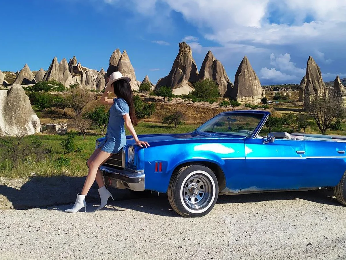 Woman posing beside a blue vintage convertible among Cappadocia fairy chimneys