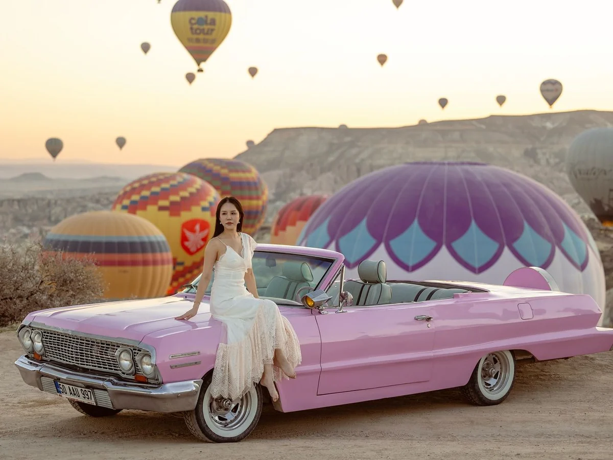 Woman posing beside a pink classic car with hot air balloons in Cappadocia