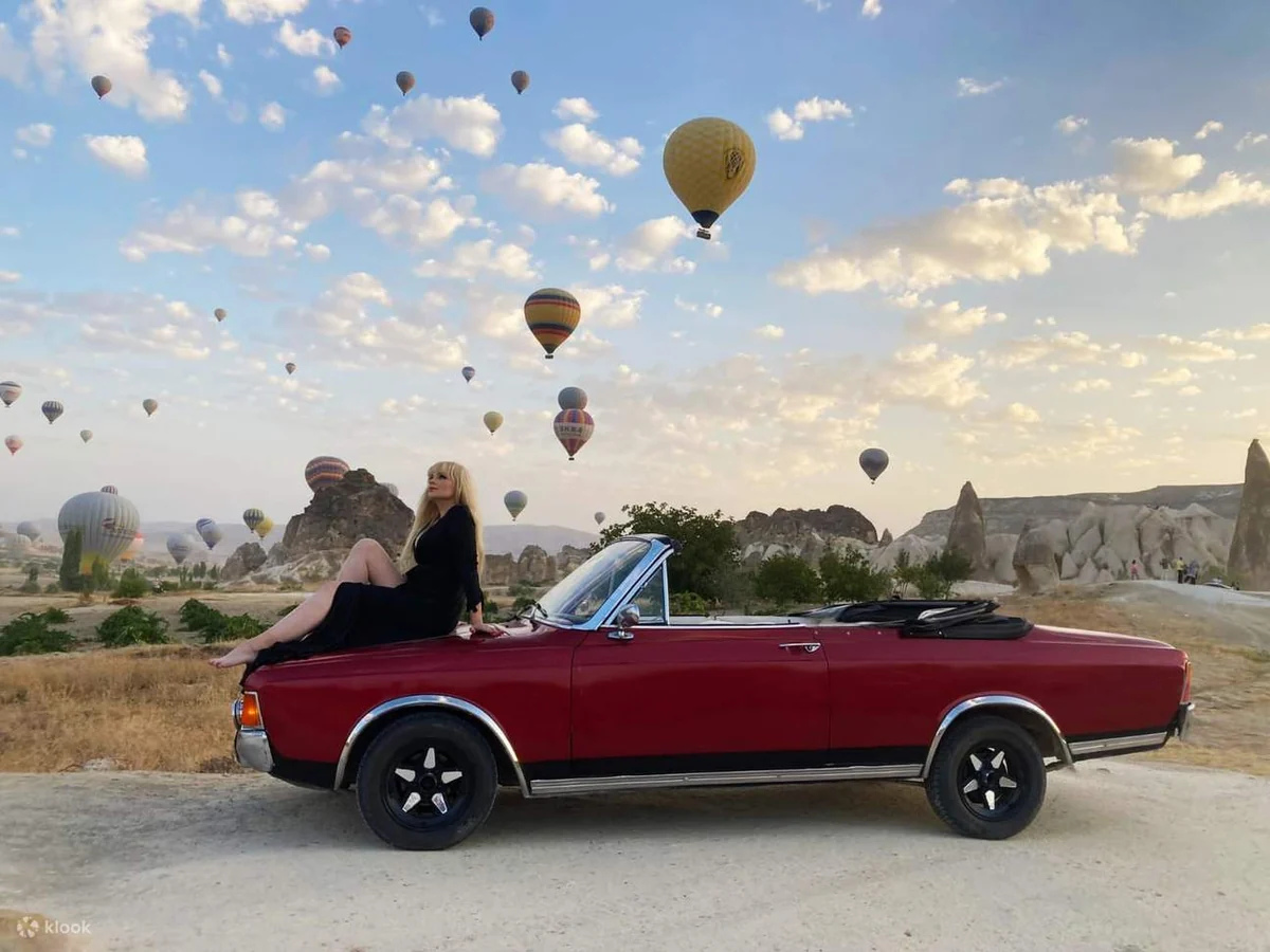 Couple seated in a red classic convertible beneath hot air balloons in Cappadocia