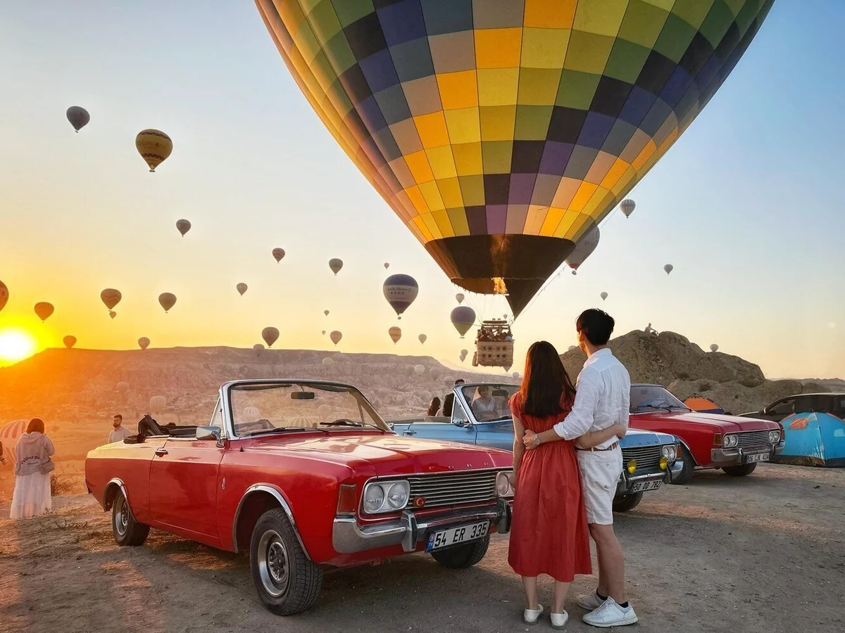 Couple beside a red vintage convertible under a large hot air balloon in Cappadocia