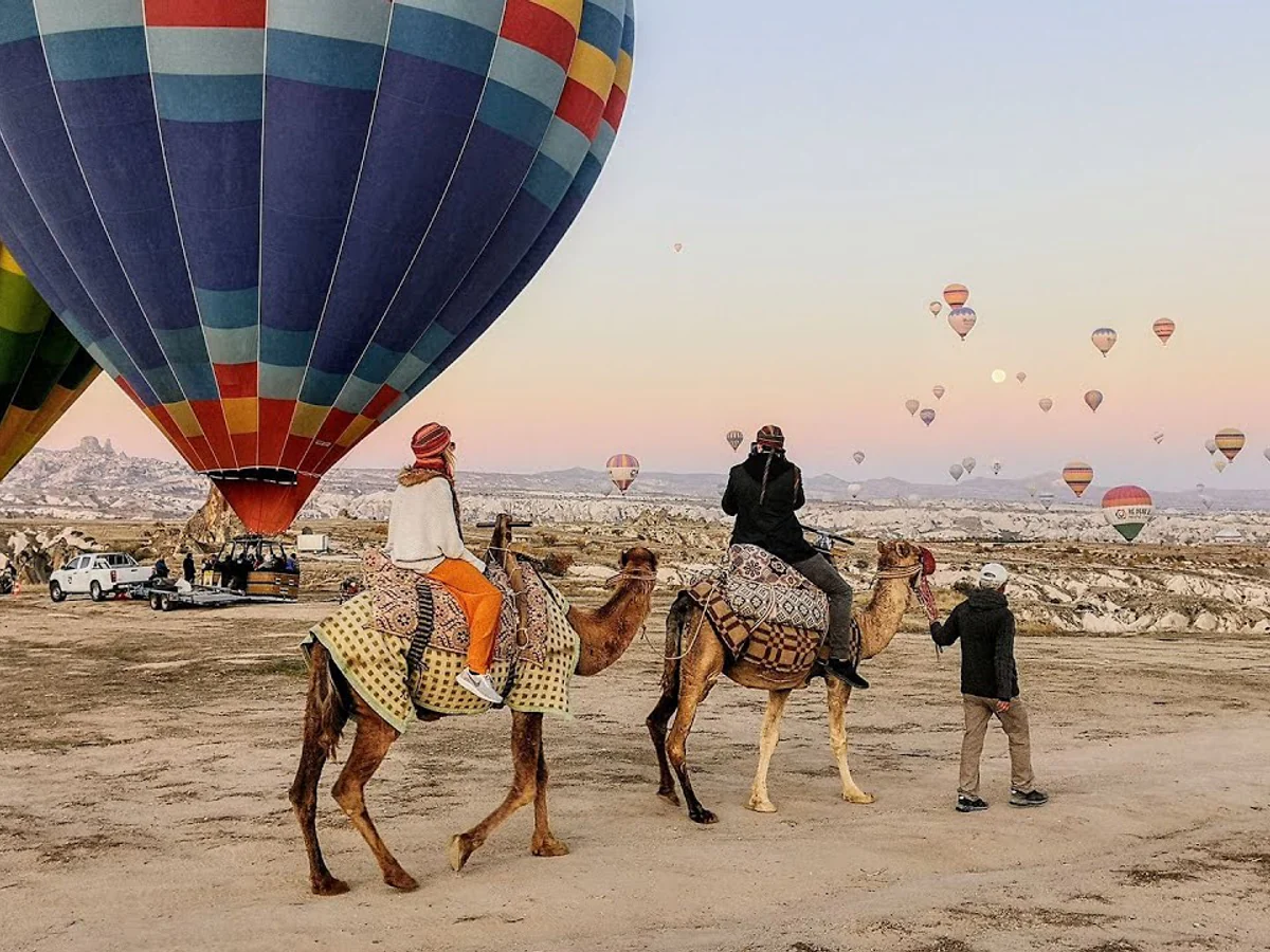Travellers riding camels beside colorful hot air balloons at sunrise in Cappadocia